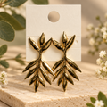 Gold-tone leaf branch earrings on cream card with botanical backdrop